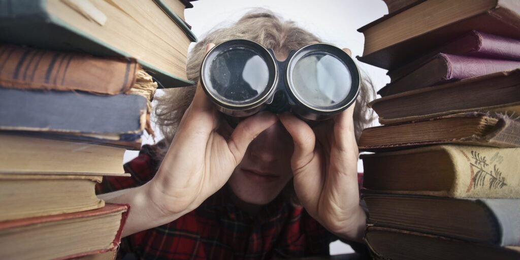 Person's head looking through binoculars with stacks of books on either side