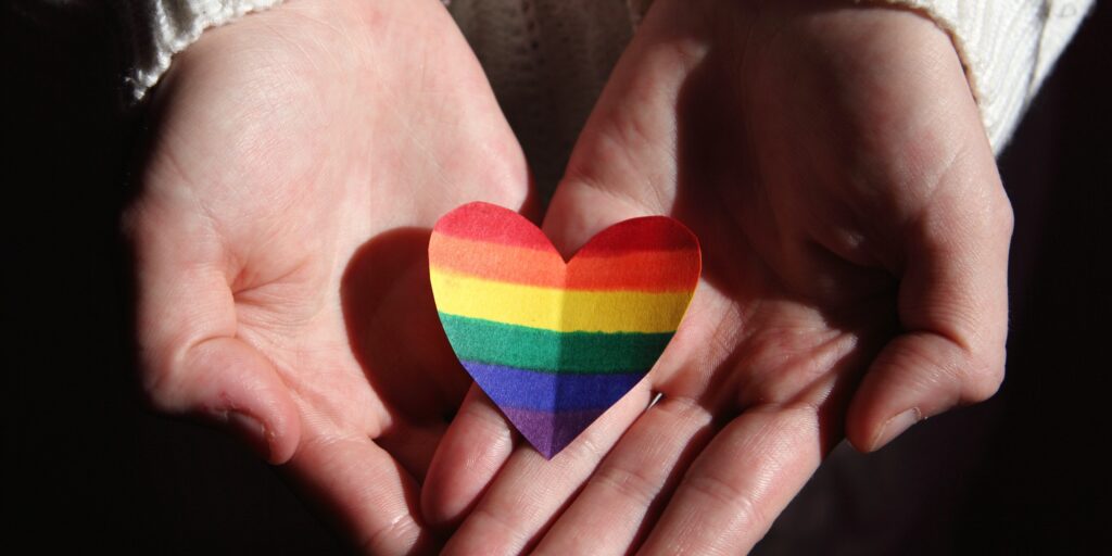 Dark background with hands holding paper heart in pride colours