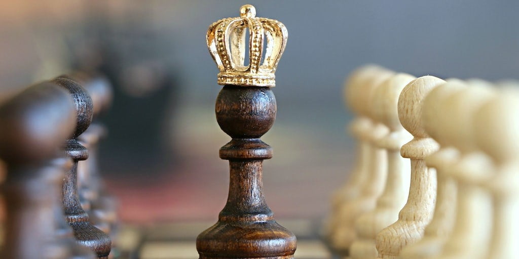 Crown on top of chess piece with more chess pieces blurred in the background.