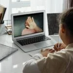 Young girl facing away from camera towards laptop where parent is shown on screen.