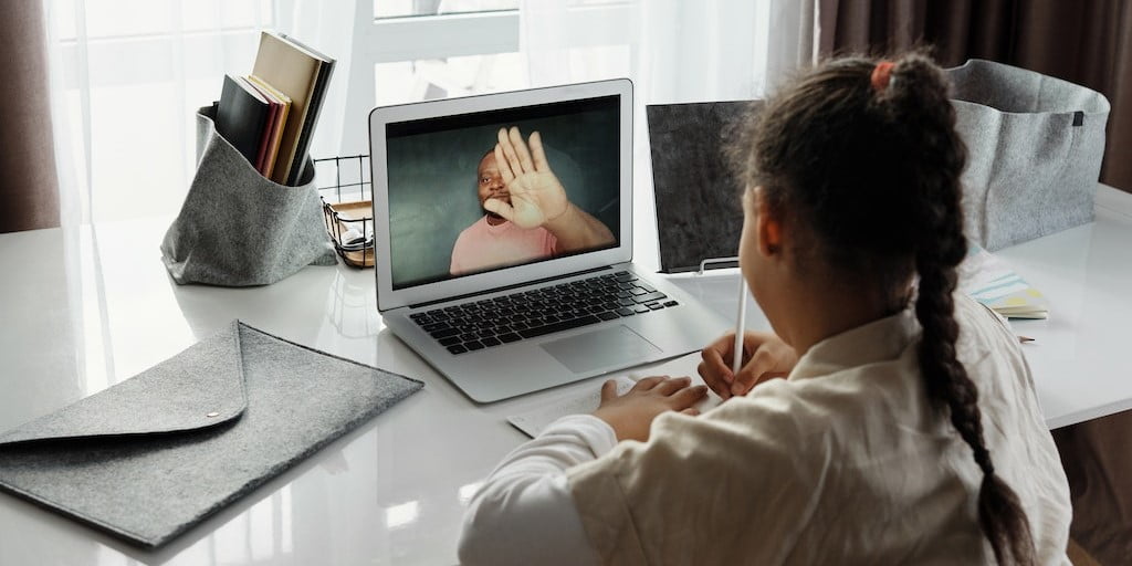 Young girl facing away from camera towards laptop where parent is shown on screen. 