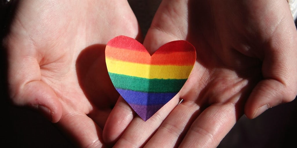 Two hands holding a heart with LGBTQ rainbow coloured stripes on it.