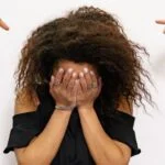 Girl standing with hands over her face and multiple fingers pointing at her on either side of her.
