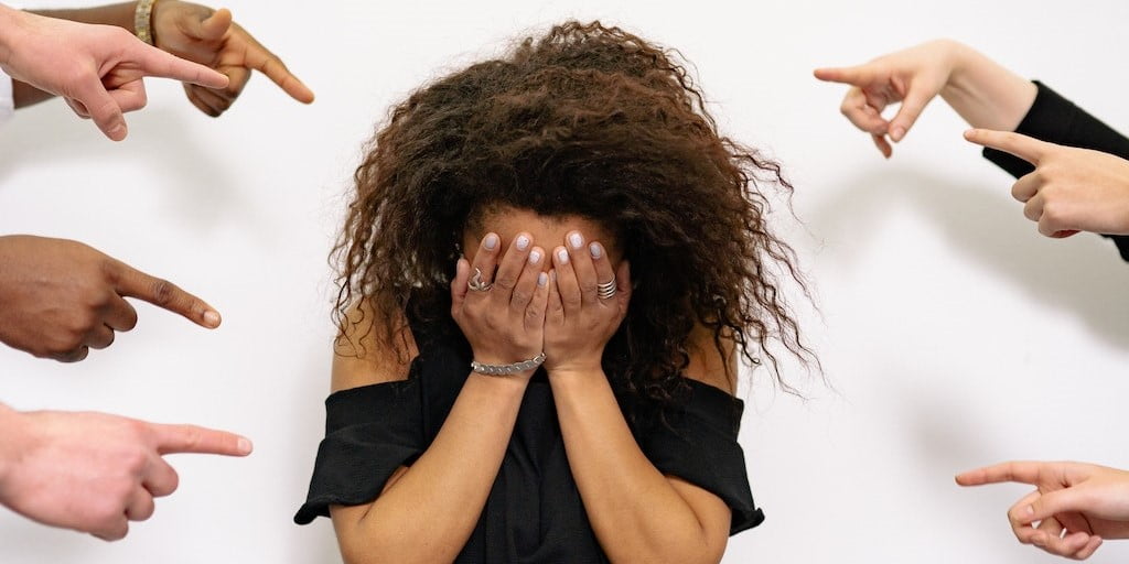 Girl standing with hands over her face and multiple fingers pointing at her on either side of her. 
