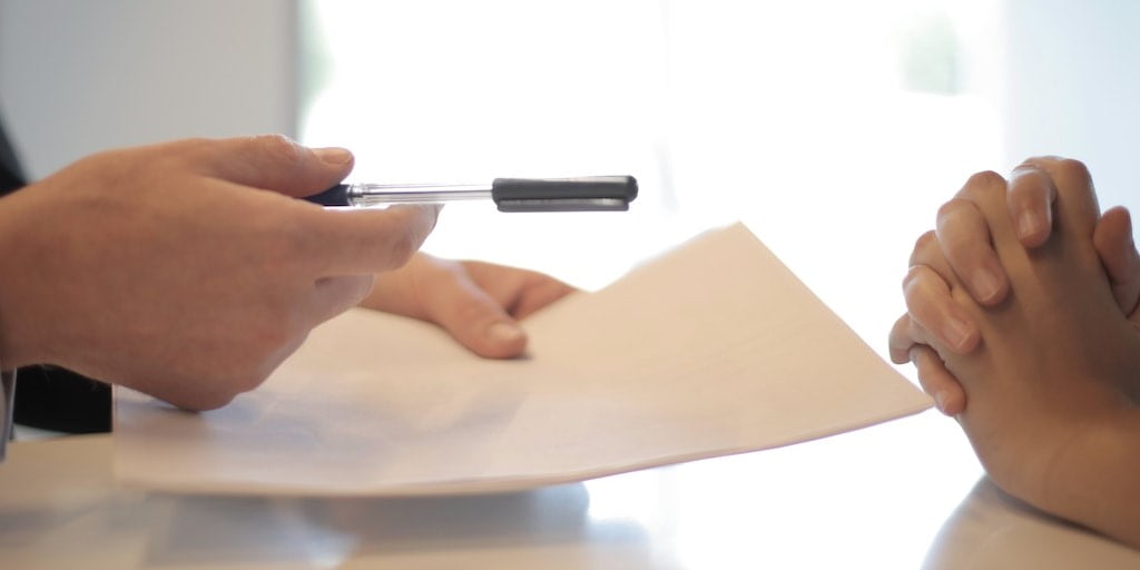 A person's hand extending a pen to another person to sign a document