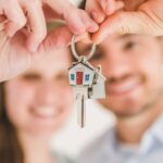 Hands holding keys in the foreground with blurred first-time home buyers in the background