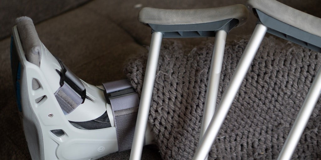 Close up of leg in walking cast, partially covered in a blanket, with crutches laying on top. 