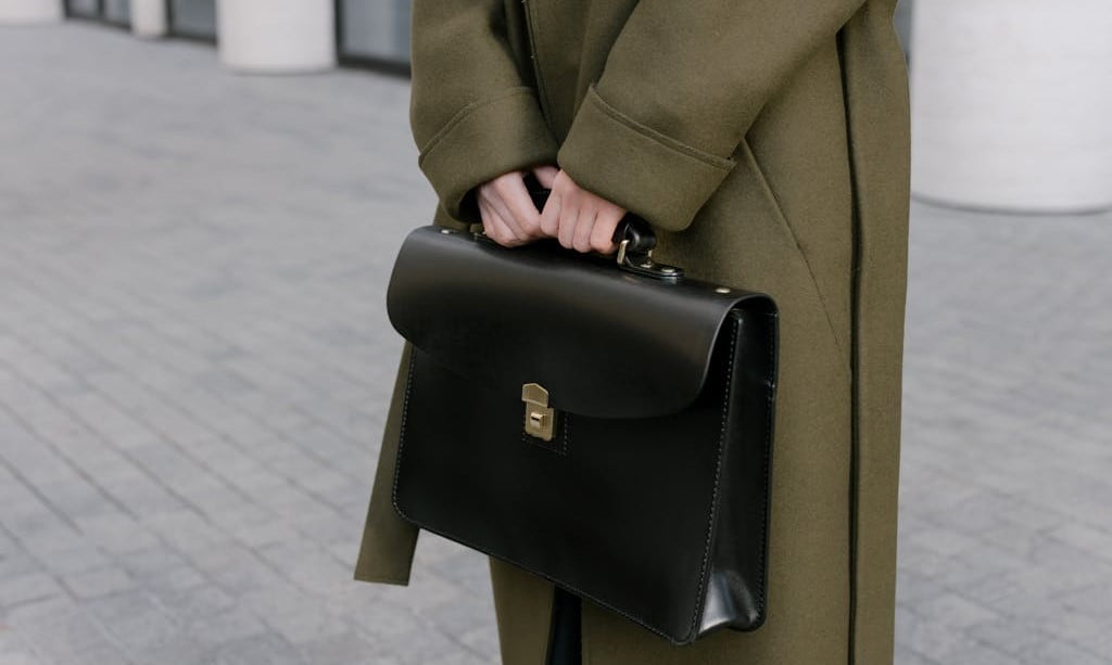 View of a person's midsection only, wearing a long green coat and holding a black leather briefcase, standing outside on grey cobblestone.