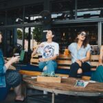 Four young women sitting outside at a cafe chatting. laughing and drinking beverages.