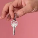 Hand holding a single key on a key ring, against a pink background