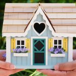 Two people's hands balancing a miniature wooden house between them.
