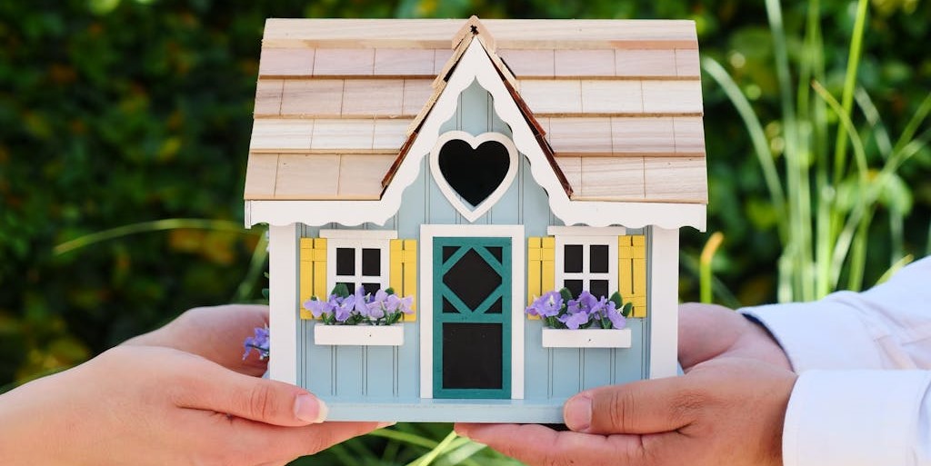Two people's hands balancing a miniature wooden house between them.