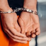 Close up view of hands in handcuffs, with orange pants and jail bars in the background