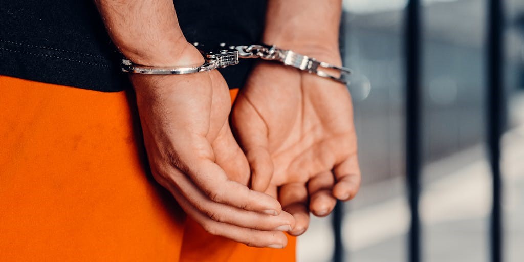 Close up view of hands in handcuffs, with orange pants and jail bars in the background