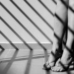 Black and white photo of legs in high heels with artistic shadow patterns on the floor. Mood is somber.