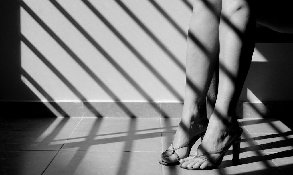 Black and white photo of legs in high heels with artistic shadow patterns on the floor. Mood is somber.
