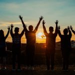 Silhouette of 7 people facing away from the camera and with their hands up with a sunset in the background.
