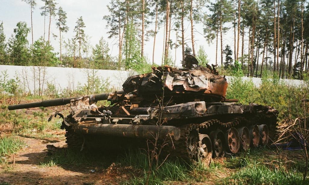 Military tank sitting damaged in grass, brush and trees.