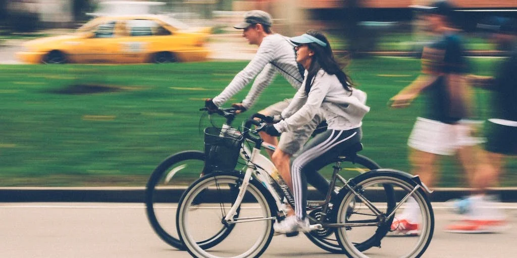 Two cyclists riding on a city street, with a blurred background of grass, cars and pedestrians.