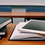 Textbooks piled on a desk beside with an open textbook with a tablet on top