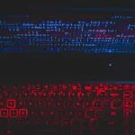 Red glow from keyboard and blue text on computer screen, all against a black background.