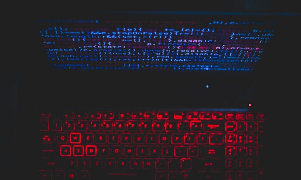 Red glow from keyboard and blue text on computer screen, all against a black background.