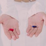 Close up of a person's hands, both open with pills in them. There's a red pill in one hand and a blue pill in the other hand.