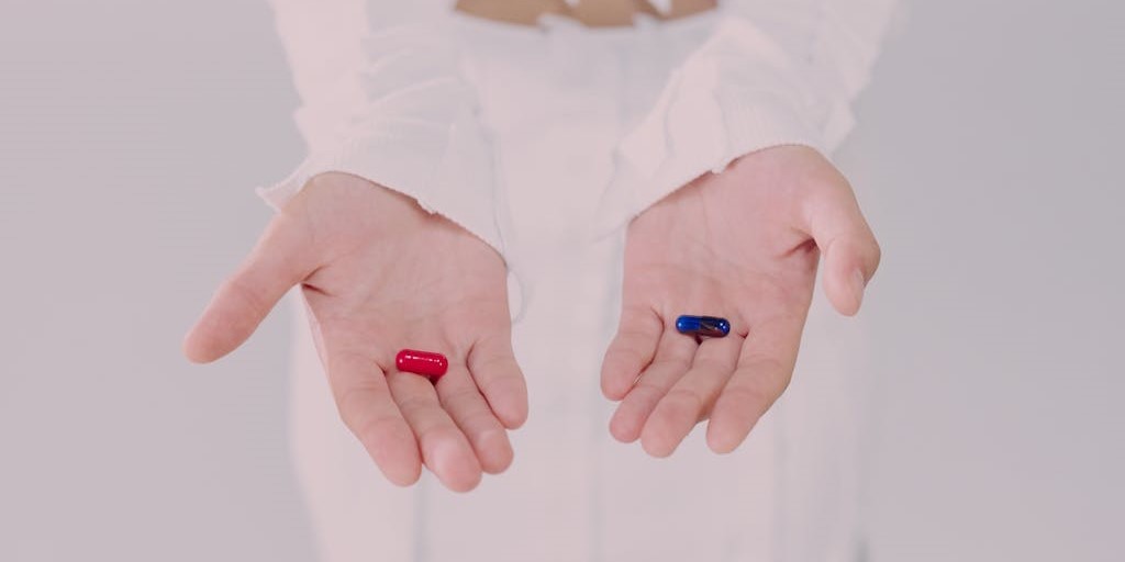Close up of a person's hands, both open with pills in them. There's a red pill in one hand and a blue pill in the other hand.
