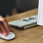 Person's hand on a computer mouse, with a view of part of a computer and keyboard in the background.